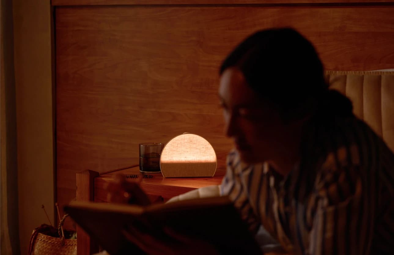 Woman reading with a lit up Hatch Sleep Clock behind her